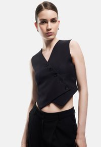 Czarny bezrękawnik o kroju typu wrap, z dekoltem w serek, czterema guzikami i dopasowanym fasonem, noszony na czarnych spodniach z wysokim stanem.