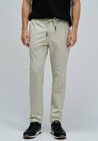 Pantalons jogger beige avec une taille élastique, fermeture à cordon, et poches latérales, associés à des chaussures de sport noires. Texture de tissu lisse.