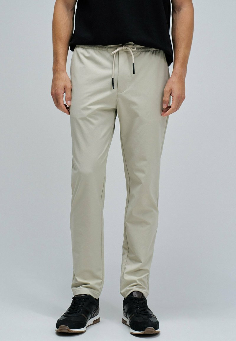 Pantalons jogger beige avec une taille élastique, fermeture à cordon, et poches latérales, associés à des chaussures de sport noires. Texture de tissu lisse.