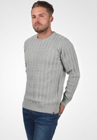 Pull en tricot gris avec un motif de torsades texturé, encolure ronde et poignets et ourlet côtelés. Porté avec un jean bleu affichant une coupe décontractée.