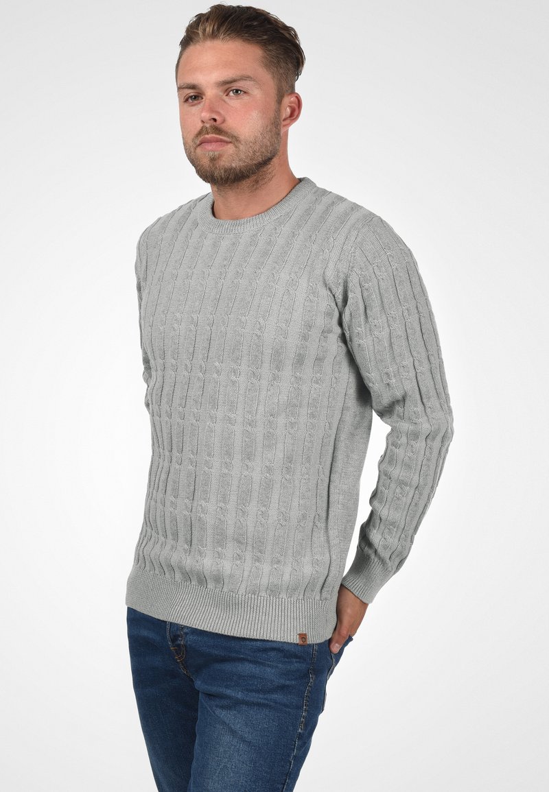 Pull en tricot gris avec un motif de torsades texturé, encolure ronde et poignets et ourlet côtelés. Porté avec un jean bleu affichant une coupe décontractée.