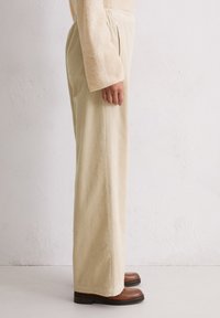 Beige Cordhosen mit weit geschnittenem Beinschnitt, ausgestattet mit vertikaler Rippstruktur und hoher Taille. Braune Lederschuhe vervollständigen das Outfit.