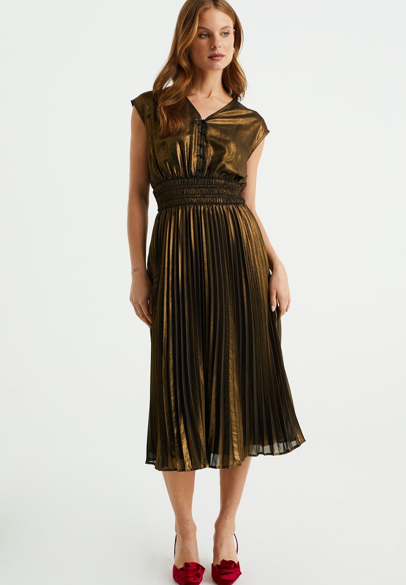 Metallisch gouden geplooide jurk met korte mouwen, elastische tailleband en V-hals; gedragen met rode hakken met strikaccenten.