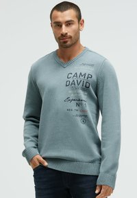 Hellblauer, strukturiert gestrickter Pullover mit V-Ausschnitt, versehen mit dunkelblauem Textdruck sowie gerippten Bündchen und Saum.