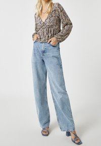 Bluse mit Blumenmuster und tiefem V-Ausschnitt, geraffter Taille und langen Ärmeln, kombiniert mit hochtaillierten, weit geschnittenen, hellblauen Jeans und blauen Absätzen.