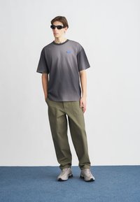 Γκρι πουκάμισο με κοντά μανίκια και διαβάθμιση χρωμάτων, με μπλε λογότυπο, συνδυασμένο με χακί παντελόνι. Φορεμένο με γκρι αθλητικά παπούτσια και μαύρα γυαλιά ηλίου, πάνω σε απλό φόντο.