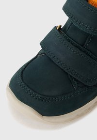 Grüner Wildlederschuh mit zwei Klettverschlüssen, runder Spitze und leichter Gummisohle. Das Innere ist mit einem kontrastierenden orangenen Stofffutter ausgestattet.