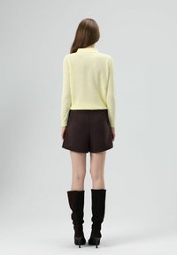 Gebreide gele trui met ruitpatroon, bruine shorts en hoge zwarte suède laarzen met een puntige hak, gezien van achteren.