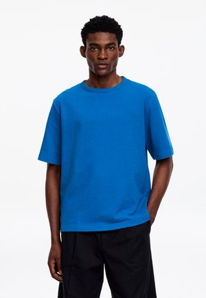 Jeune homme debout, une main dans la poche, portant une chemise unie bleu vif à manches courtes et un pantalon noir sur fond blanc.
