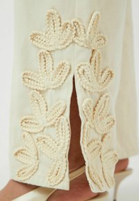 Pantalones de color crema con bordados decorativos en un patrón de hojas a lo largo del lado, y un detalle de abertura en el dobladillo para un diseño adicional.