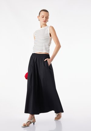 Mujer con top sin mangas a rayas blanco, falda larga negra, sandalias blancas de tacón, sosteniendo un bolso de mano rojo, posando sobre fondo blanco.