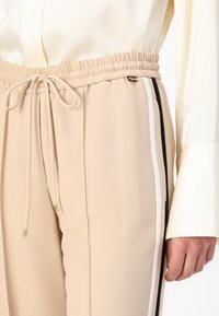 Beige joggingbroek met een elastische tailleband, trekkoorddetail en zijstrepen in zwart en wit. Textuurstof zichtbaar.