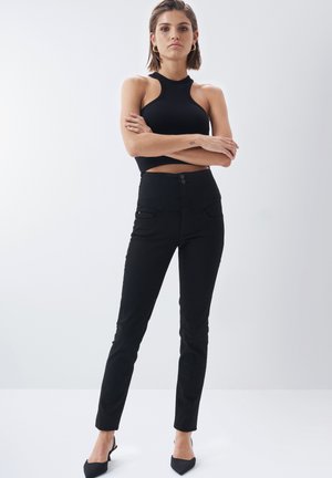 Jeans negros de talle alto con bolsillos frontales, cierre de dos botones y diseño ajustado. Combinados con una blusa sin mangas de canalé negra.