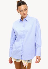 Mujer vestida con una camisa de botones oversized de color azul claro con una pequeña "L" bordada en blanco y pantalones cortos con estampado, de pie contra un fondo blanco.