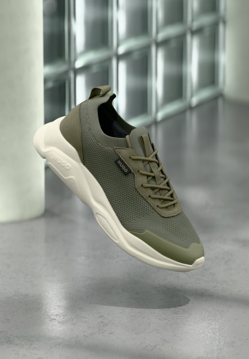Baskets tricotées vert olive avec semelle blanche épaisse et étiquette "HUGO", flottant au-dessus d'un sol gris poli près de briques en verre.
