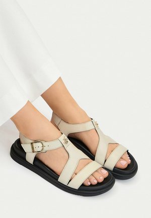 Piedi che indossano sandali in pelle beige chiaro con suole nere e cinturini con fibbia dorata su sfondo bianco.