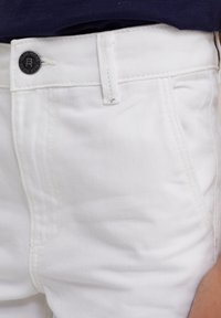 Witte spijkerbroek met een klassiek vijf-pocketontwerp, een knoopsluiting met een logo en subtiele textuurstoffen. Schone stiksels accentueren.