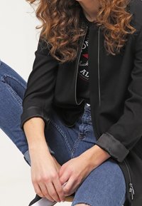 Chaqueta negra con cremallera y textura suave, de mangas largas, combinada con jeans de mezclilla azules. La chaqueta presenta un diseño sin costuras y sutiles acentos metálicos.