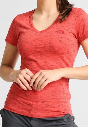 Kobieta ubrana w dopasowaną czerwoną koszulkę z krótkim rękawem i dekoltem w serek, z delikatnymi czarnymi cętkami oraz małym logo na piersi, z lekko splecionymi dłońmi.