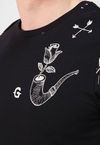 Camisa negra con un gráfico blanco de una pipa humeante sosteniendo una rosa en flor, flechas cruzadas arriba y una pequeña letra "G" en el pecho.