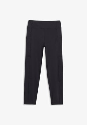 Jeune personne debout portant un t-shirt graphique rose, des leggings noirs et des chaussures de randonnée noires sur un fond clair uni.