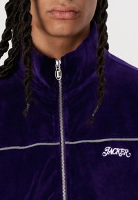 Lilla zip-up jakke laget av myk stoff, med grå horisontale striper, og "JACKER" logo brodert i hvitt på brystet.