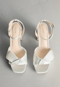 Biele saténové sandále na vysokom podpätku s ozdobou v tvare mašle, štvorcovou špičkou, členkovými remienkami a hladkým, lesklým povrchom. Názov značky "NEXT" je na vnútorných vložkách.