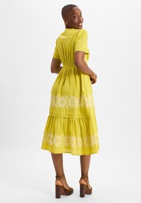 Yellow midi dress made of lightweight fabric, featuring a smocked waist, short sleeves, and white floral embroidery along the hem. Brown heeled sandals.