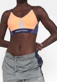 El sujetador deportivo Nike Pro presenta paneles en rosa y azul marino, una franja central gris y tirantes ajustables. El material es elástico y ceñido.