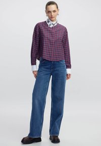 Camicetta a quadri rossi e blu, corta con maniche lunghe e colletto bianco; abbinata a jeans larghi a vita alta blu e scarpe scure.