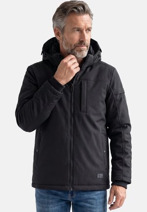 Mittelalter Mann mit grauem Haar und Bart, trägt eine schwarze Kapuzenjacke mit Reißverschluss und blaue Jeans, richtet den Jackenkragen.