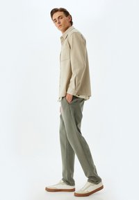 Camisa de botones beige con bolsillo frontal, combinada con pantalones verde ajustados y zapatillas blancas con suelas marrones.
