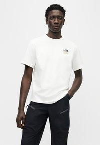 Άνδρας που φοράει λευκό κοντομάνικο T-shirt με μικρό λογότυπο στο στήθος και μαύρο παντελόνι με τσέπες με φερμουάρ, στέκεται μπροστά σε ανοιχτόχρωμο φόντο.