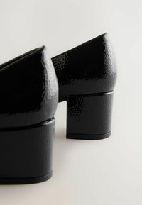 Black patent leather block-heeled shoes with a pointed toe and textured finish, showcasing a smooth surface and thick heel design.
