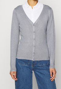 Cárdigan gris con escote en V, mangas largas, cierre de botones y cintura ajustada. Lleva puesto sobre una camisa blanca con cuello y jeans azules.