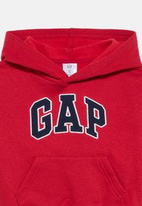 Červená dětská mikina s kapucí, klokáním kapsou a velkým tmavě modrým logem "GAP" orámovaným bílou barvou na přední straně.