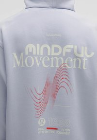 Lyslilla hættejakke med "MINDFUL Movement" trykt i hvid, som har et rødt bølgedesign og teksten "1998 FIND FLOW WITHIN THE JOURNEY".