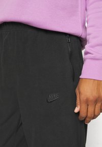 Pantaloni jogger neri in pile con tasca con zip, logo Nike ricamato e cintura elastica; abbinati a una maglietta a maniche lunghe di un leggero viola.