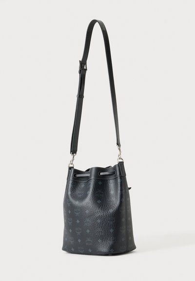 Sac seau noir texturé avec logos subtilement imprimés en motif et une longue bandoulière réglable sur un fond clair uni.