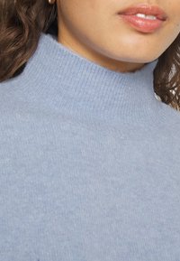 Ljusblå polotröja med ett ribbat mönster, med en hög halsdesign och mjukt stickat tyg, delvis synlig i bilden.