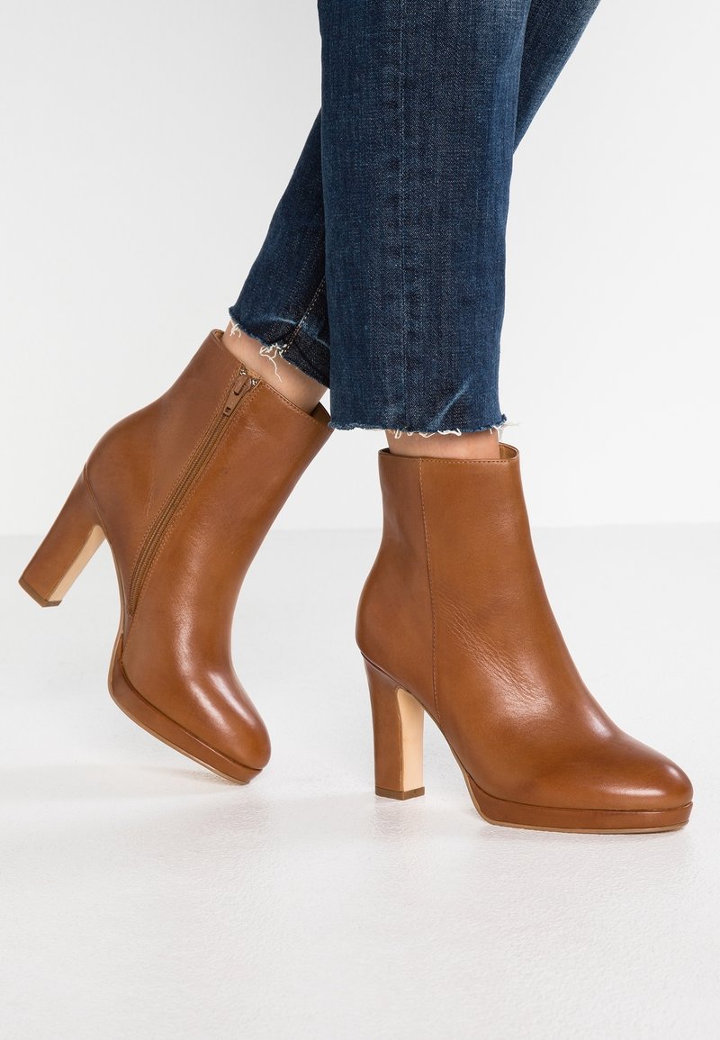 Bottines en cuir marron avec fermetures éclair latérales, portées avec un jean bleu foncé à ourlet effiloché sur fond blanc.