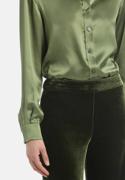 Zelena satenska bluzica s gumbima sprijeda i dugim rukavima, u kombinaciji s tamnozelenim baršunastim hlačama koje imaju uski pojas i mekani tekstur.