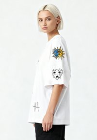 Balta medvilninė per didelė marškinė su juodais ir spalvingais siuvinėtais raštais, vaizduojančiais saulę ir mėnulį, bei širdį su veido bruožais ant rankovių.