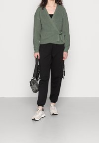 Person klädd i en grön omslagströja, svarta cargobyxor, vita sneakers, hållande en axelväska med leopardmönster mot en enfärgad bakgrund.
