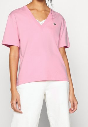 Femme portant un haut rose à manches courtes en V avec un petit logo vert sur la poitrine et un pantalon blanc sur un fond uni.