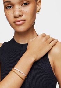 Mujer con piel con pecas que lleva una camiseta sin mangas negra, pulseras de oro y perlas, y pendientes de oro y perlas a juego.