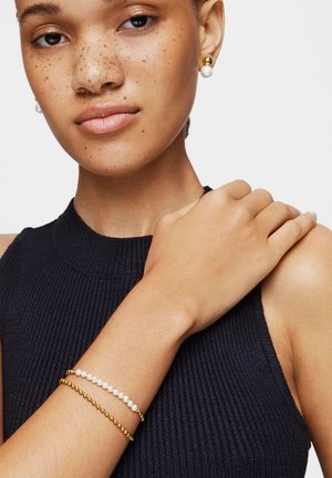 Donna con pelle lentigginosa che indossa un top nero senza maniche, braccialetti d'oro e perle e orecchini a perno abbinati d'oro e perle.