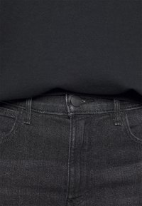 Sudadera de algodón negra que cubre parcialmente unos pantalones vaqueros negros de talle alto. Los pantalones presentan una textura sutil y un cierre de botón metálico.