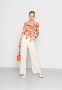 Top cropped de manga longa com um padrão de espiral vermelho e laranja, combinado com calças largas cor creme. Os acessórios incluem uma pequena carteira laranja.