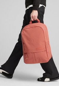 Zaino corallo con forma arrotondata, tessuto texture, maniglia superiore per il trasporto e tasca con pattina frontale. Abbinato a pantaloni neri e scarpe slip-on.